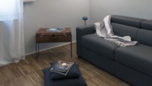 Ein modernes Wohnzimmer mit einem grauen Sofa, einem kleinen Holztisch mit einem Buch und einer Zeitschrift sowie einer blauen Stehlampe.