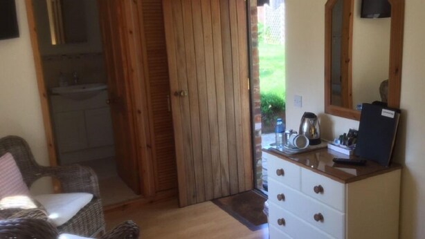 Una stanza con una porta in legno, una sedia in vimini, un comò bianco con cassetti e uno specchio appeso al muro.