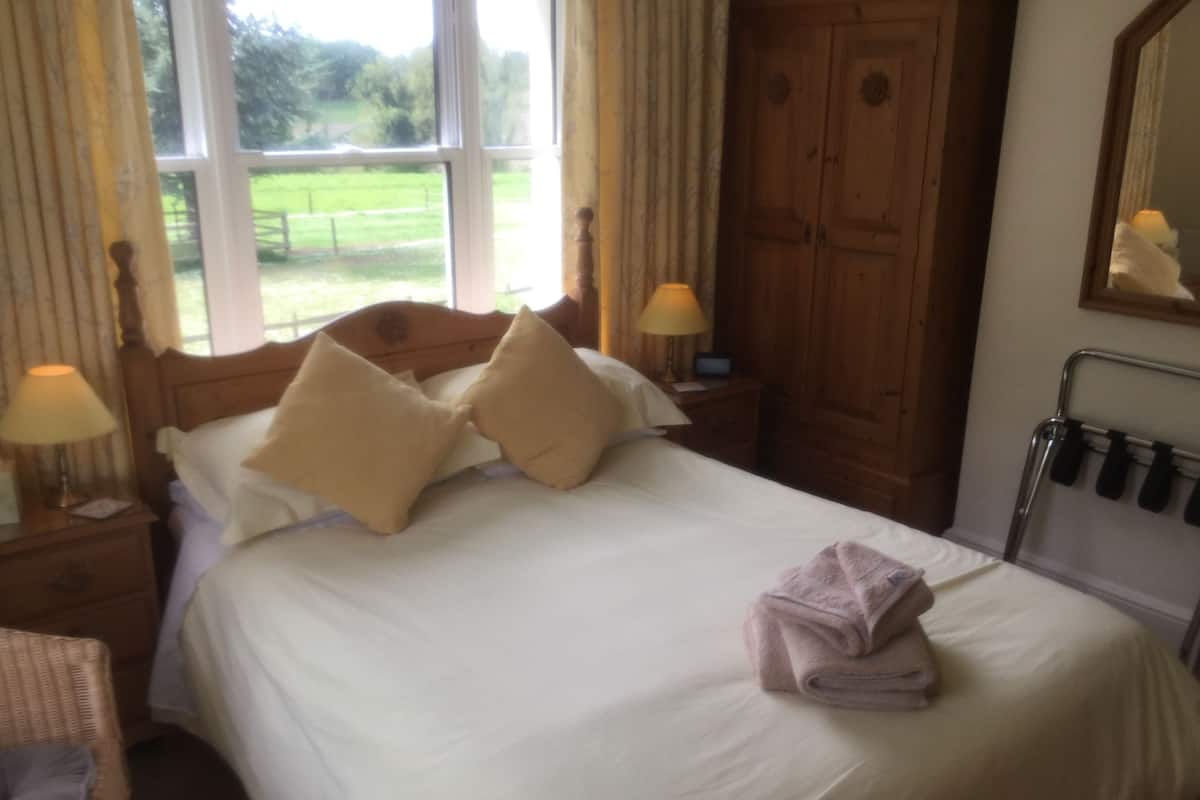 Una cama bien hecha con sábanas blancas y dos almohadas color beige, una lámpara de noche, un armario de madera y una ventana con vistas a zonas verdes.