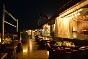 An outdoor dining area with wicker chairs and tables, potted plants, and a view of the city skyline at night. - Hotel Surya Plaza (Kota)