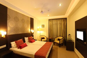 A hotel room with a large bed, two armchairs, a small table, a TV, and a ceiling fan. - Hotel Surya Plaza (Kota)