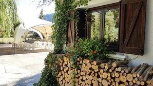 Un espacio al aire libre rústico con sillas y mesas de plástico blanco, un muro de troncos de madera y una ventana con contraventanas de madera.