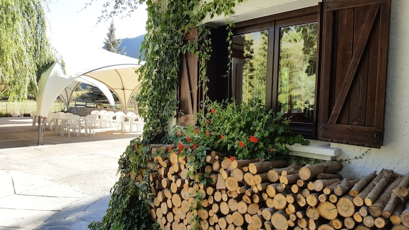 A rustic outdoor seating area with white plastic chairs and tables, a wooden log wall, and a window with wooden shutters.