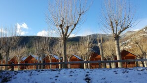 Una hilera de cabañas de madera, con el suelo cubierto de nieve y árboles sin hojas al fondo.