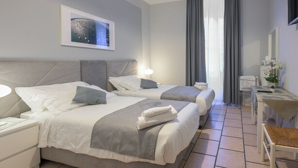 A hotel room with two beds, a bedside table, a chair, a desk, and a large window with curtains. - Affittacamere Berzi (Trieste)