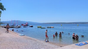 Une plage où l’on voit des gens se baigner et se promener le long du rivage, des bateaux sur l’eau et un ciel dégagé.