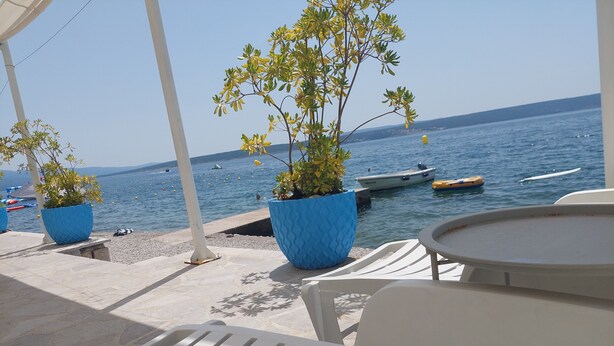 Une terrasse en bord de mer, avec des chaises blanches et des jardinières bleues, donnant sur des eaux calmes où naviguent des bateaux.