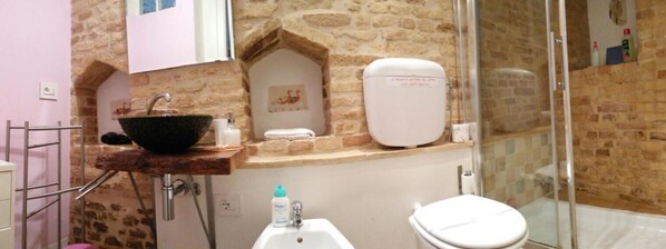 A bathroom with a stone wall, a sink with a black basin, a toilet, and a glass shower enclosure. - B&B La Lanterna (Fermo)