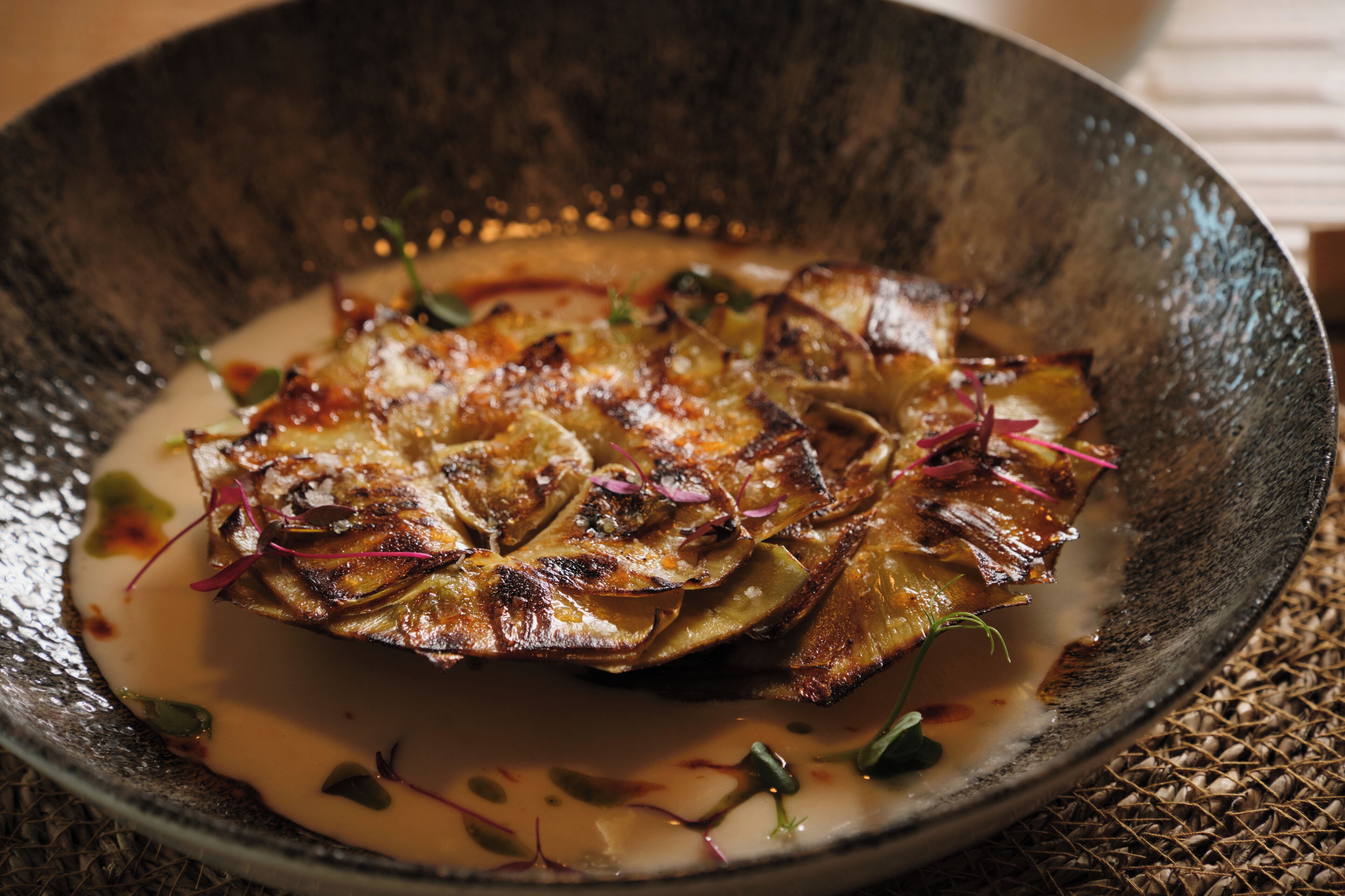 Un plato en un tazón con superficie texturizada, adornado con hierbas y flores comestibles.
