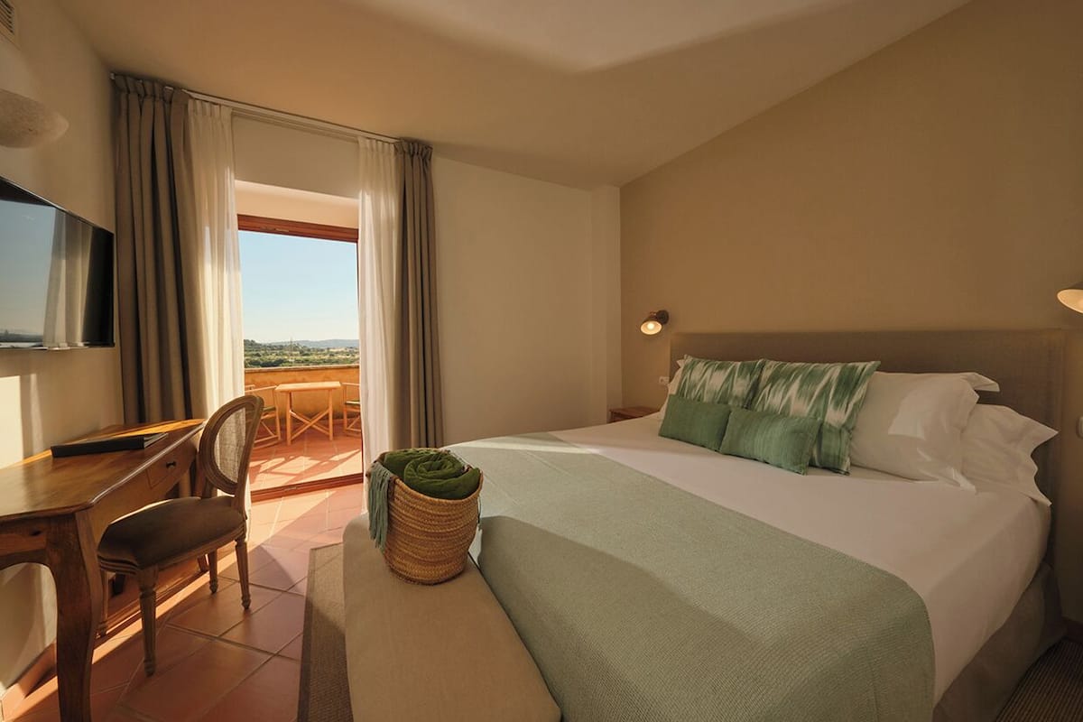 A hotel room with a bed, a desk, a chair, a TV, and a view of the outdoors.
