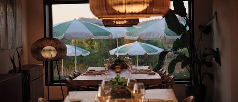 A dining area with a long table set for a meal, large umbrellas, and a view of greenery through a window.