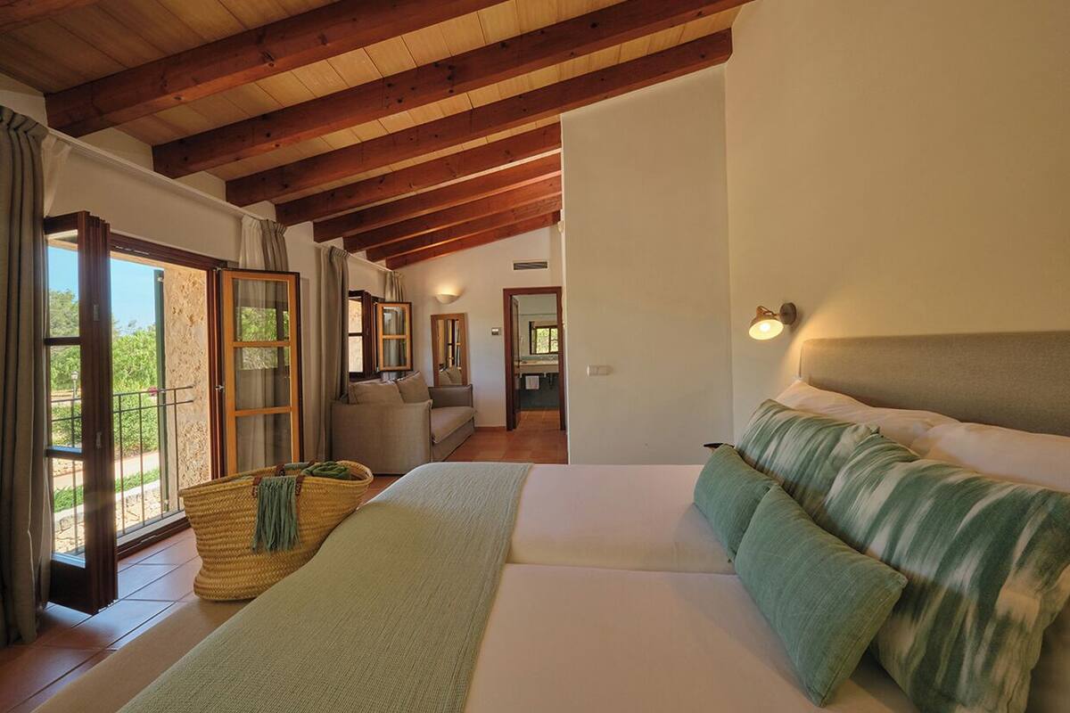 A bedroom with a large bed, wooden beams, a view through a glass door, and a small sitting area with a sofa.