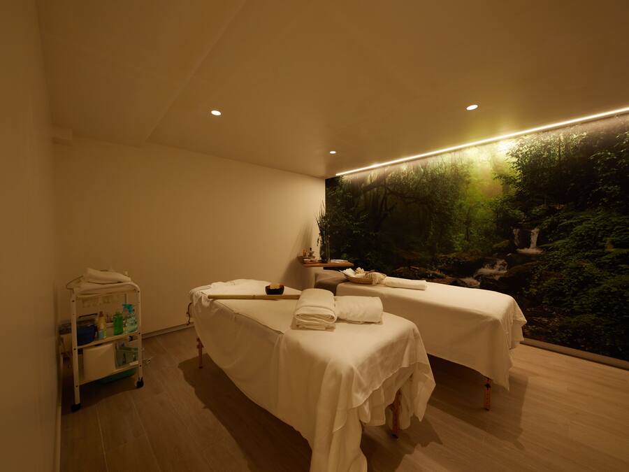 Un cuarto de spa con dos camillas de masaje, un carrito con toallas y artículos de aseo, y un gran mural en la pared que representa un bosque.