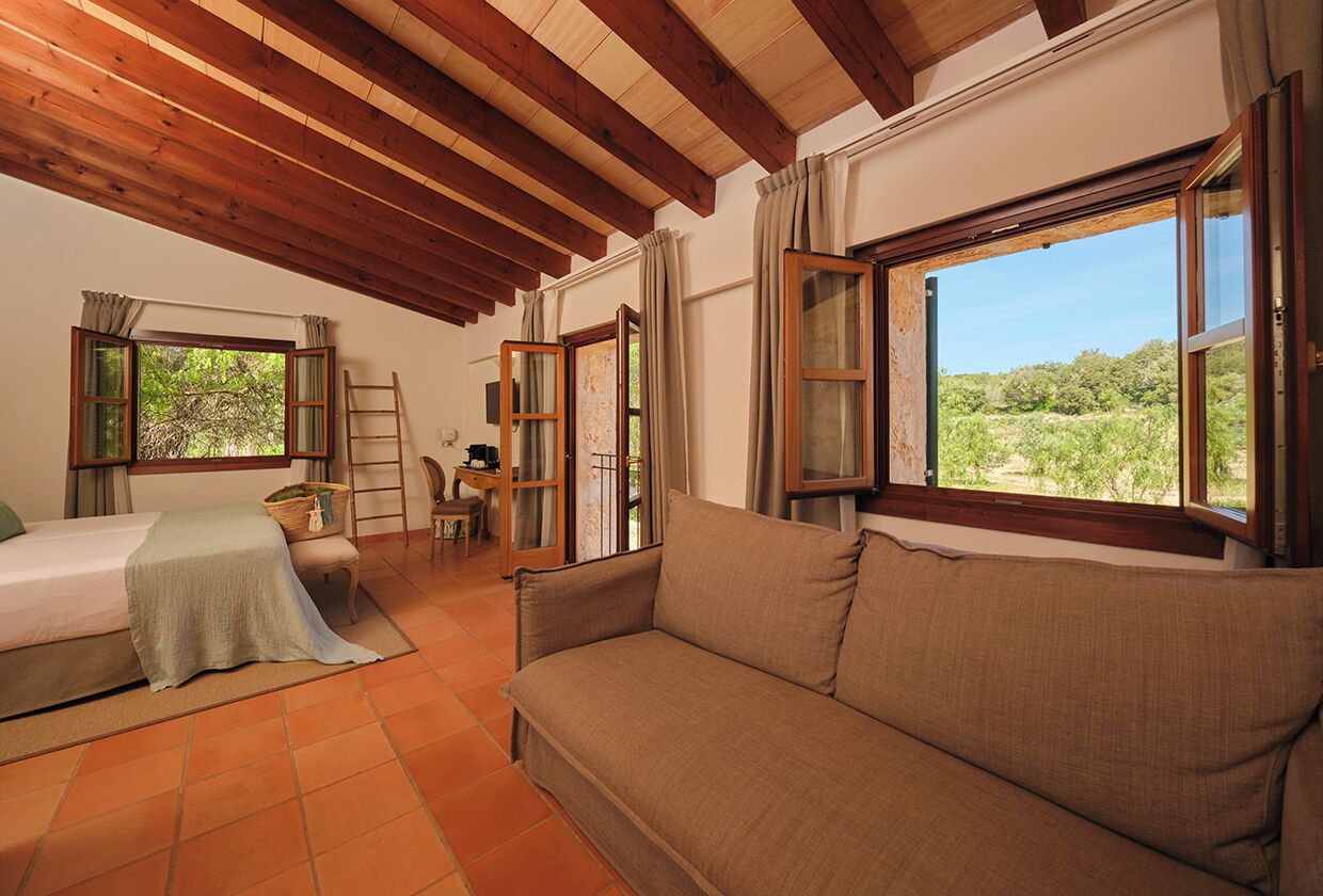 Quarto com cama, sofá, escrivaninha e cadeira, e janelas amplas com cortinas.