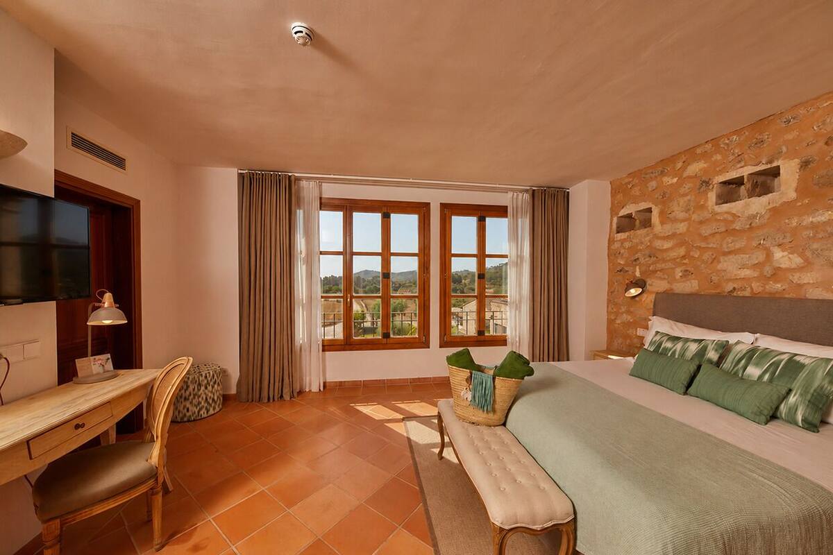 A bedroom with a bed, a desk, a chair, a window with curtains, and a stone wall.