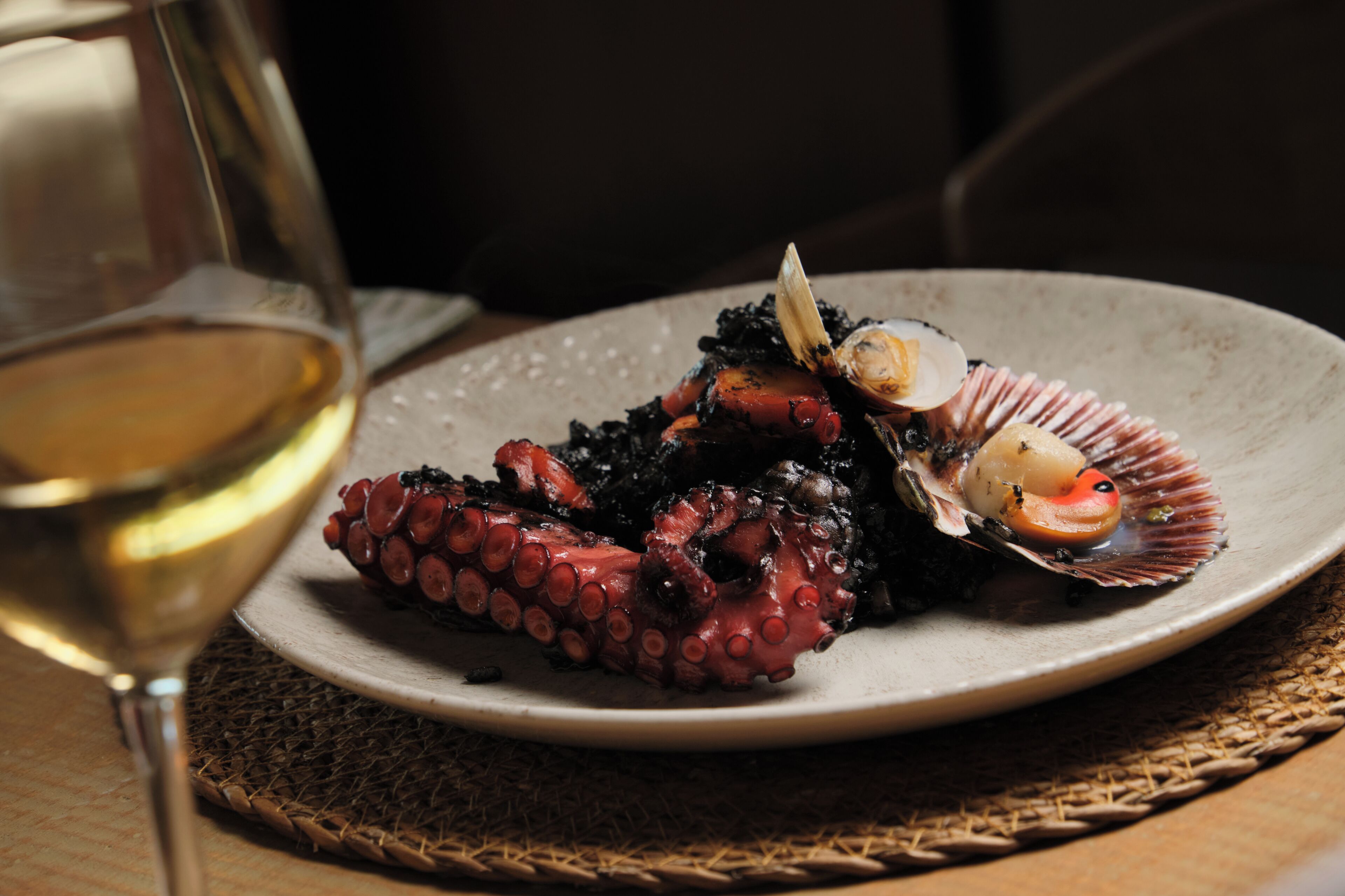 Un plato de pulpo con una concha de vieira, una copa de vino blanco y un mantel tejido.