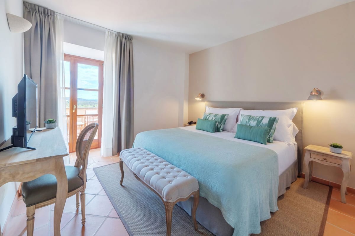 A bedroom with a bed, a desk with a chair, a bench, a nightstand, and a view of the outdoors.