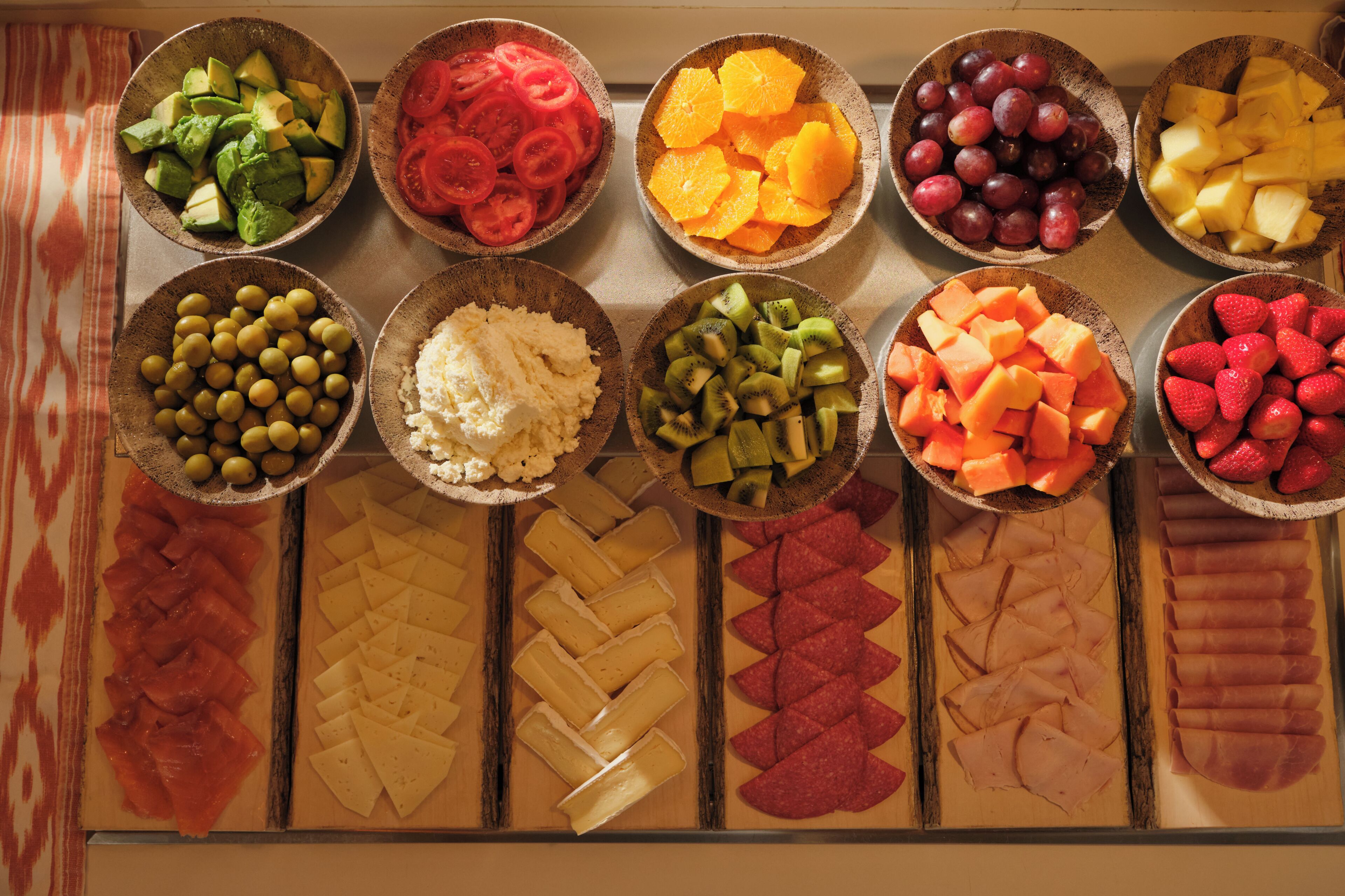Variedad de alimentos dispuestos en cuencos y una tabla de madera, incluyendo frutas, verduras, quesos y carnes.