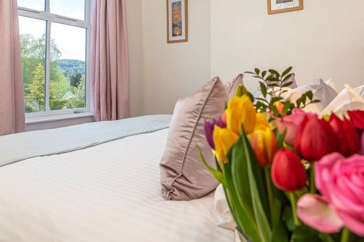 A bedroom with a bed, a window with curtains, and a bouquet of flowers.