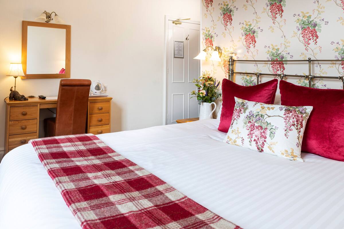 A neatly made bed with a red and white checkered blanket, a wooden desk with a chair, a mirror, and a wall with floral wallpaper.