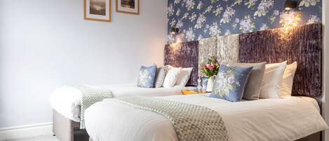 Une chambre avec deux lits, un papier peint à motifs floraux, des tableaux encadrés accrochés au mur et une tête de lit décorative.