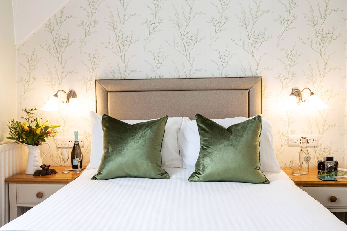 A neatly made bed with two green pillows, a nightstand with a lamp, a vase with flowers, and a bottle of wine.