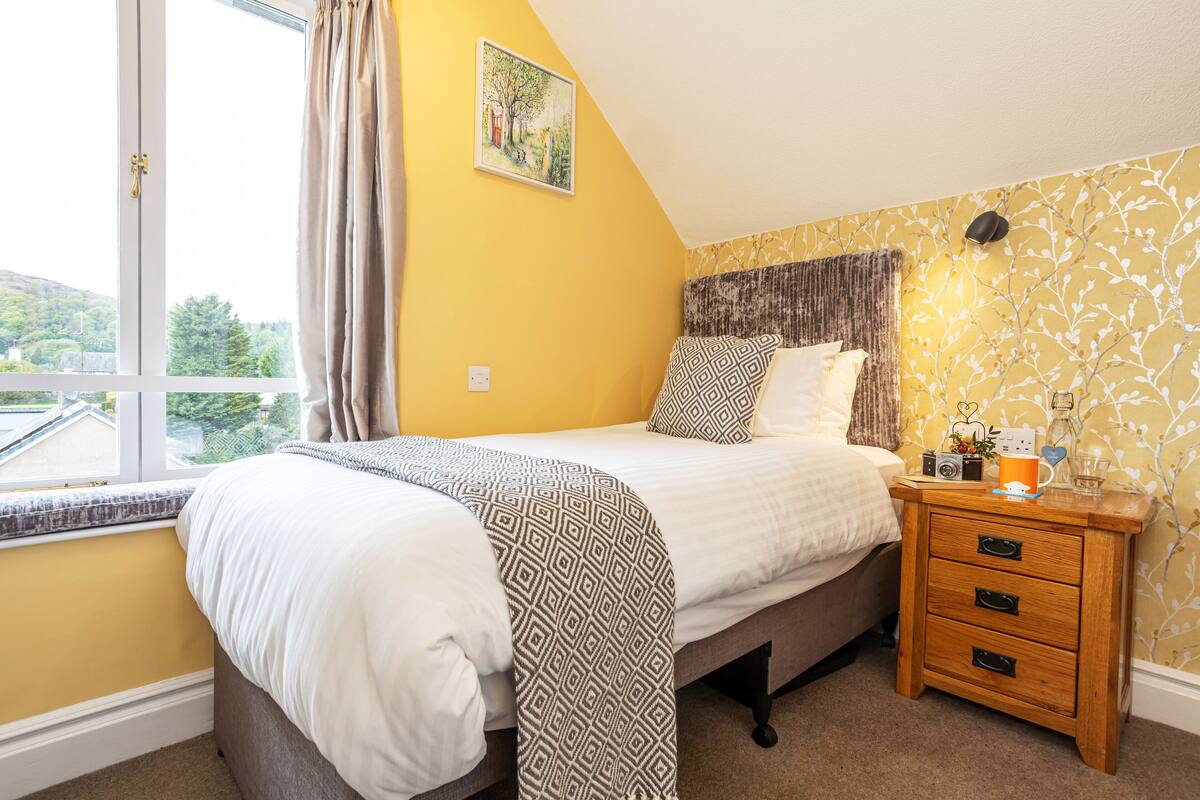 A bedroom with a bed, a bedside table with a camera, and a window with curtains.