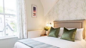 A bedroom with a large bed, a nightstand with a lamp, and a window with curtains. - Elder Grove (Ambleside)