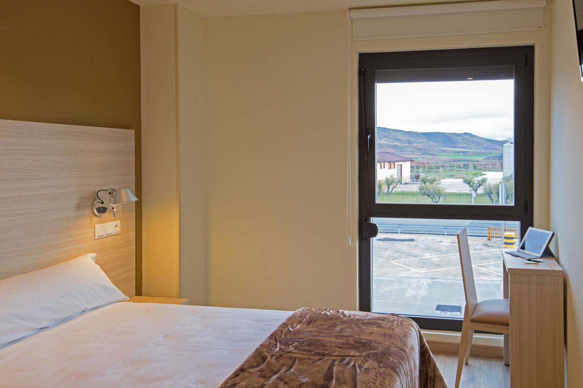 A hotel room with a bed, a desk with a laptop, and a view of the outside.