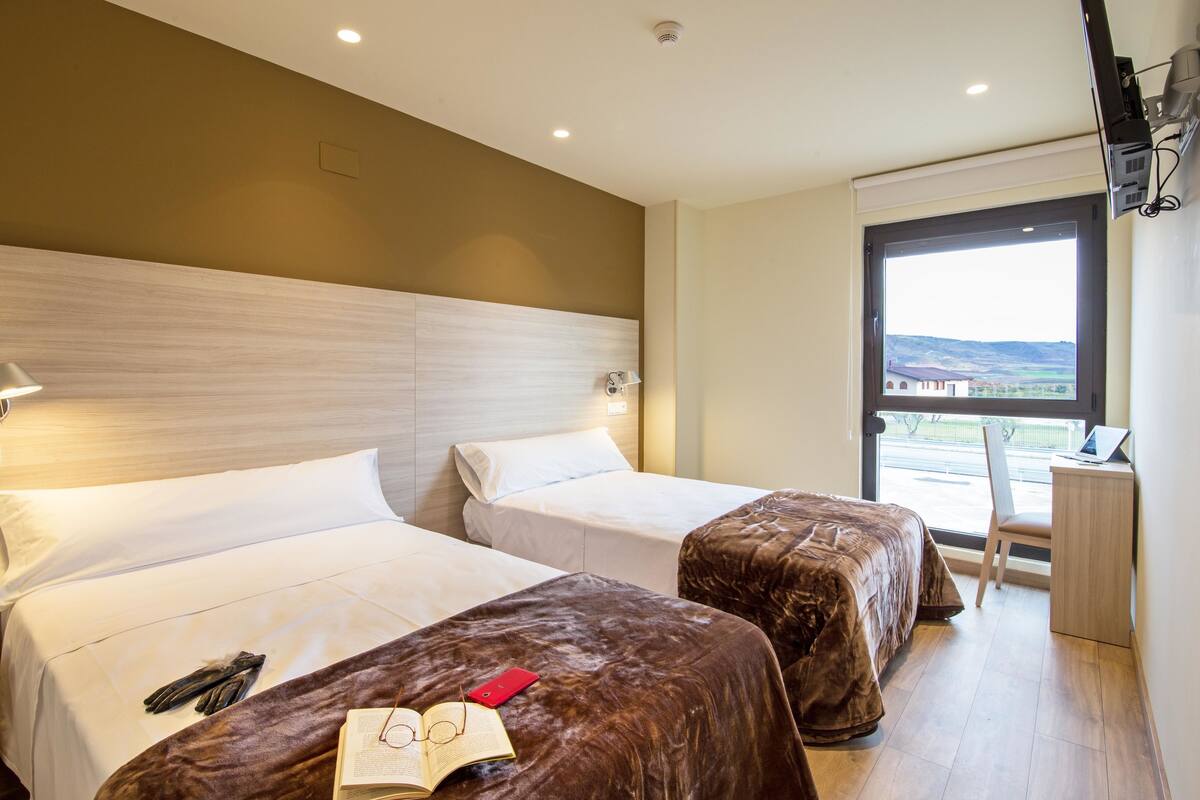 A hotel room with two beds, a desk, and a window with a view.