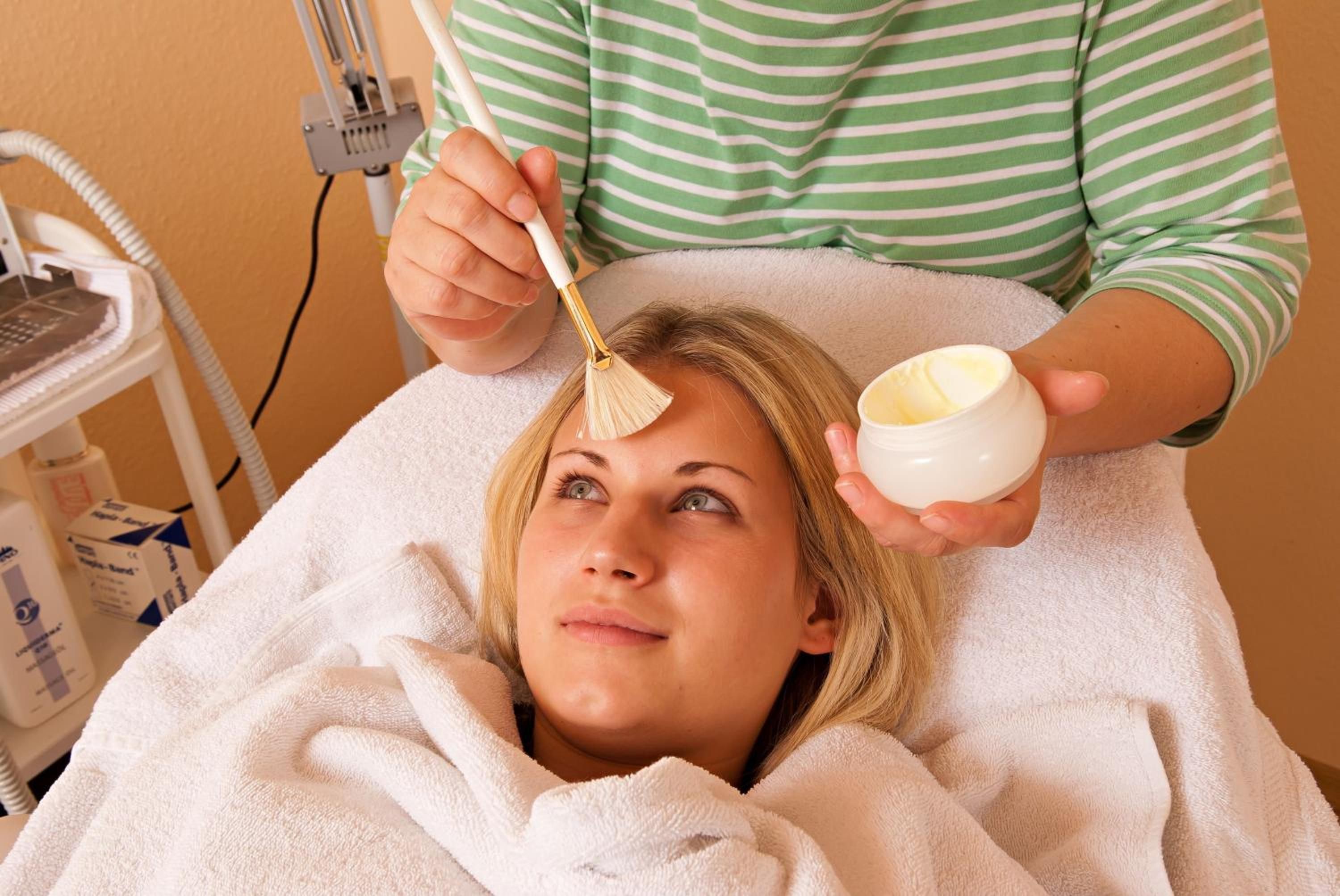 Une personne qui reçoit un soin du visage à l’aide d’un pinceau, d’un bol de crème et d’un appareil médical visible en arrière-plan.