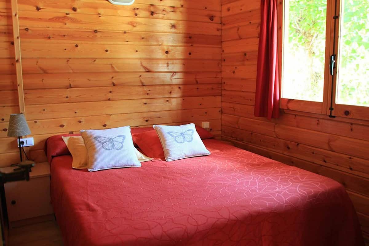 Una habitación de cabaña de madera con una cama, dos almohadas, una mesita de noche, una lámpara y un ventanal con cortinas.