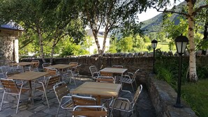 Café al aire libre con paredes de piedra, mesas de madera y sillas de metal, rodeado de árboles y un muro de piedra.