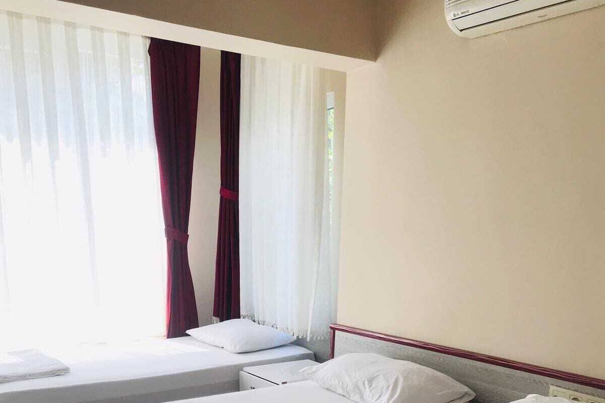 Hotellihuoneessa on kaksi erillistä sänkyä, joissa on valkoiset lakanat ja tyynyt. Huoneessa on ikkuna, jossa on läpikuultavat valkoiset verhot ja tummempi punainen verho, joka on osittain vedettynä. Ilmastointilaite on asennettu seinälle sänkyjen yläpuolelle.