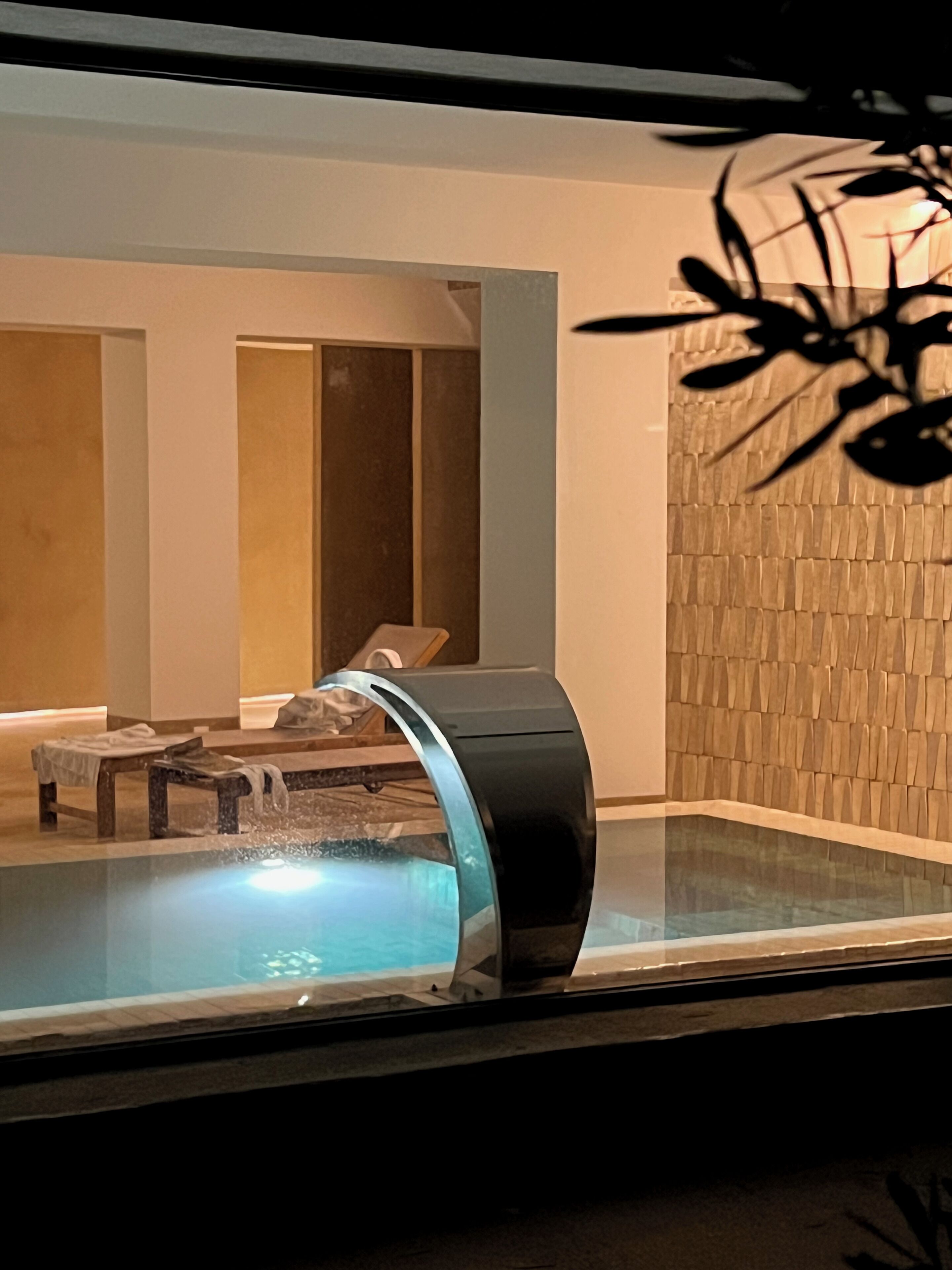 A modern indoor pool with a curved edge, a lounge area with a bench and a table, and a textured wall in the background.