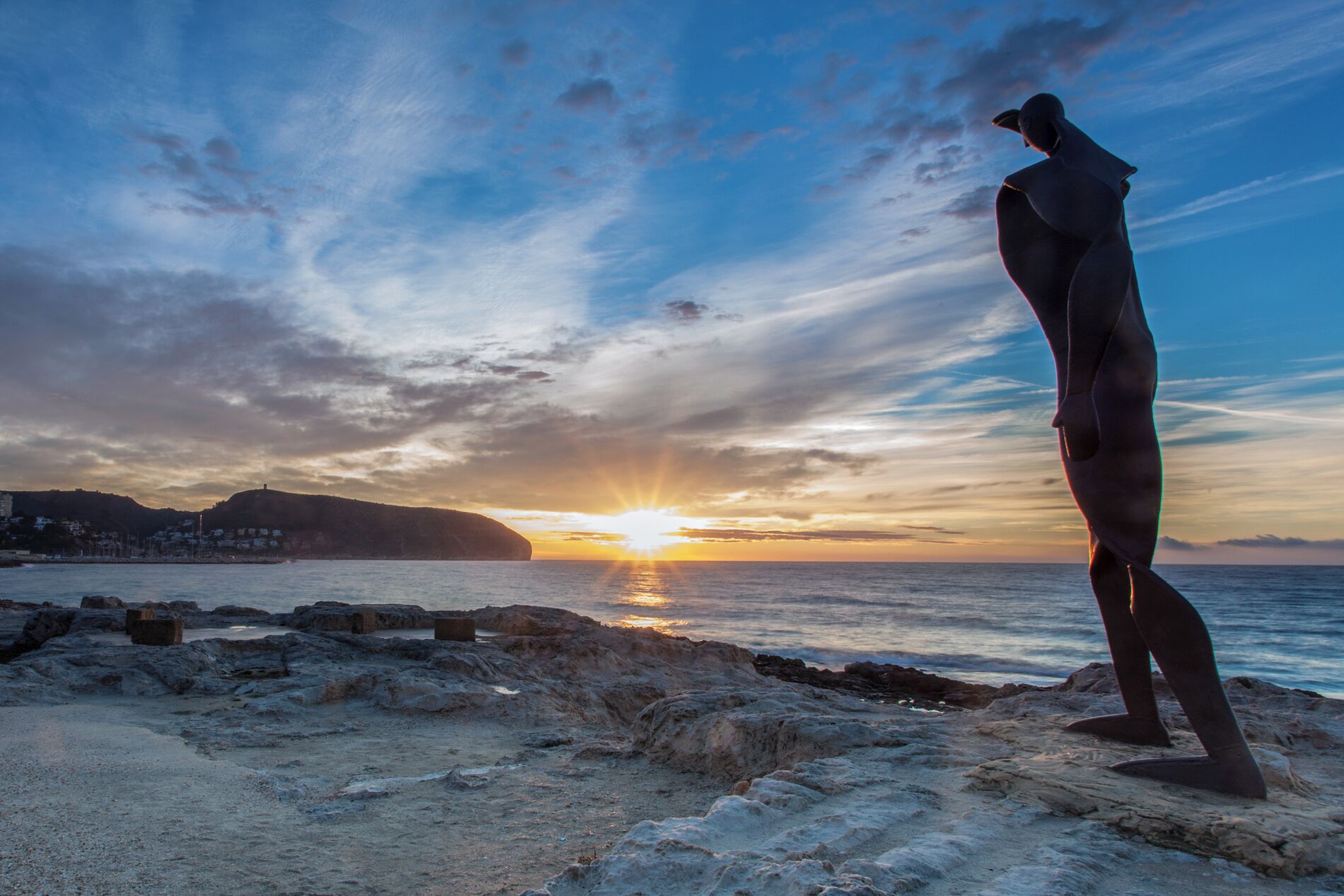 Eine Person steht an einem felsigen Strand bei Sonnenuntergang mit Blick auf das Meer und eine entfernte Hügelkette.