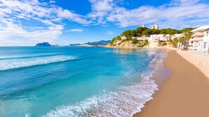 A beach with clear blue water, a sandy shore, and buildings along the coastline. - Hotel Serawa Moraira (Teulada)