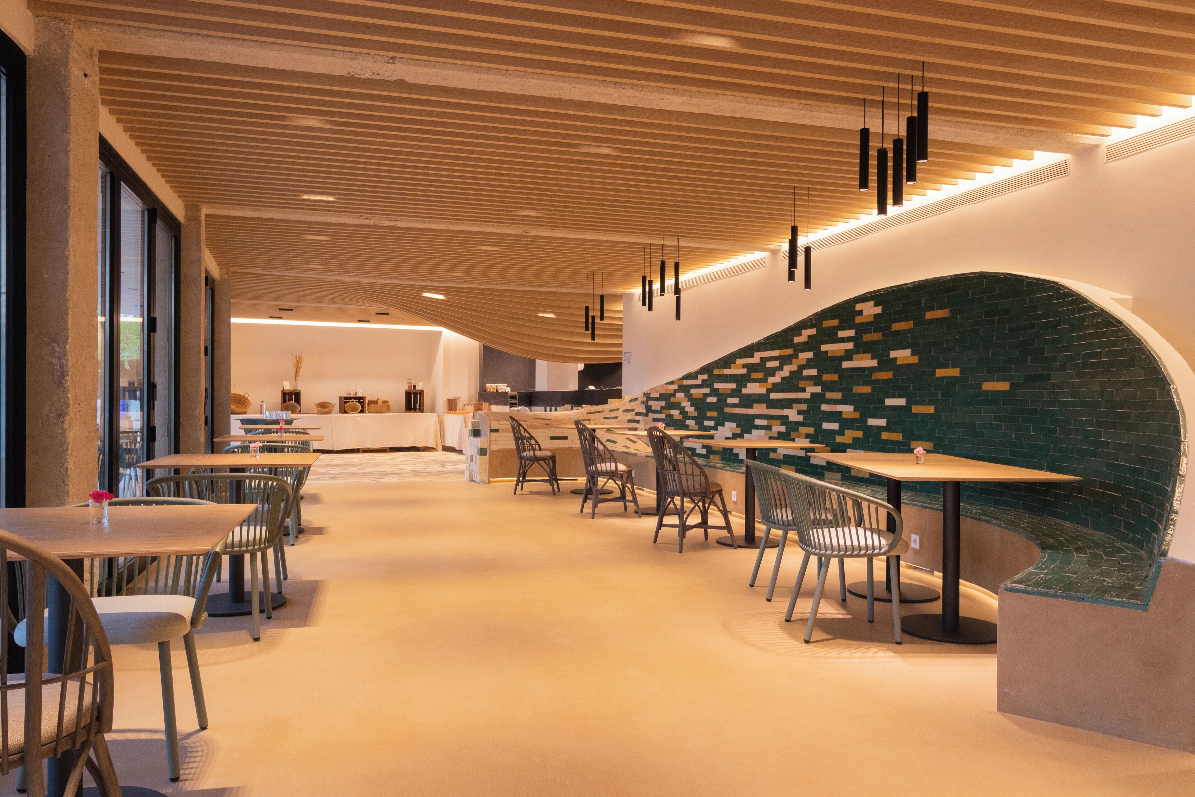 Een modern restaurantinterieur met houten plafondpanelen, zwarte hanglampen en een groene bakstenen muur. Tafels en stoelen staan klaar voor het diner.