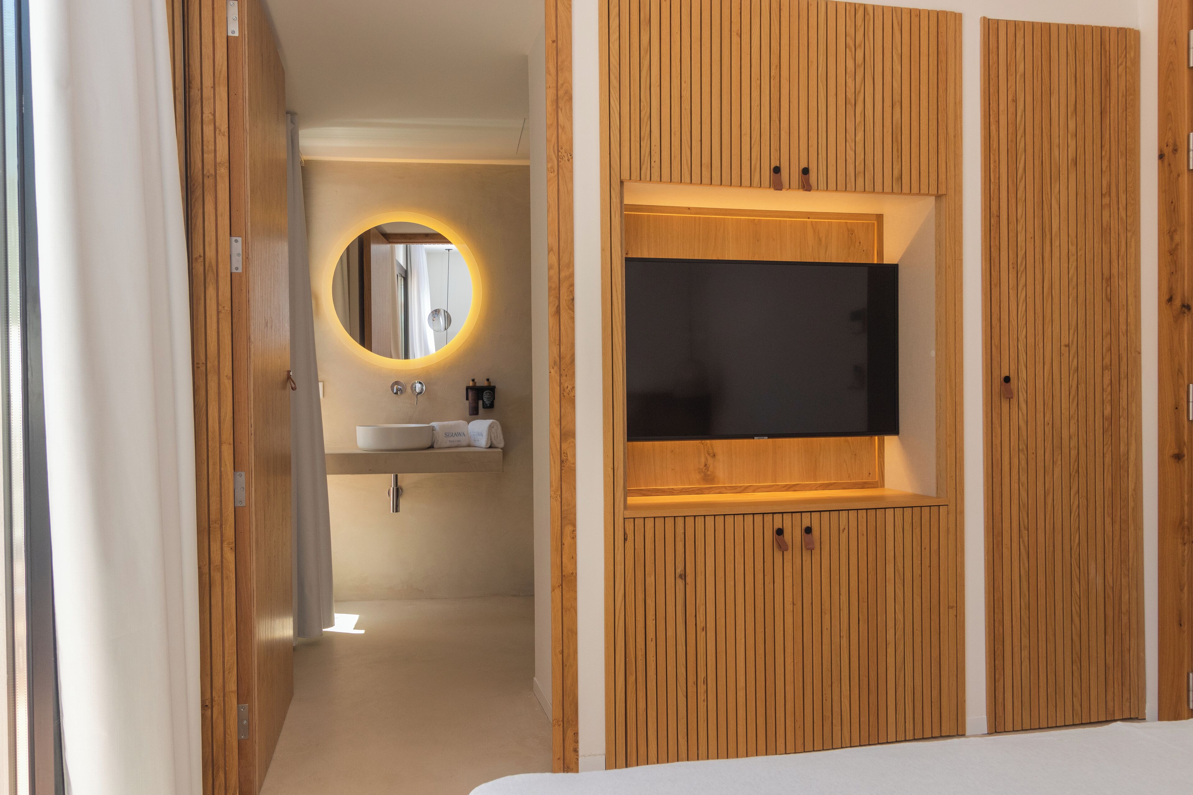 A modern bathroom with a wooden vanity, a round mirror, and a built-in TV.