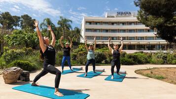 Um grupo de pessoas praticando yoga em tapetes ao ar livre, com um edifício identificado como "SERAWA" ao fundo.