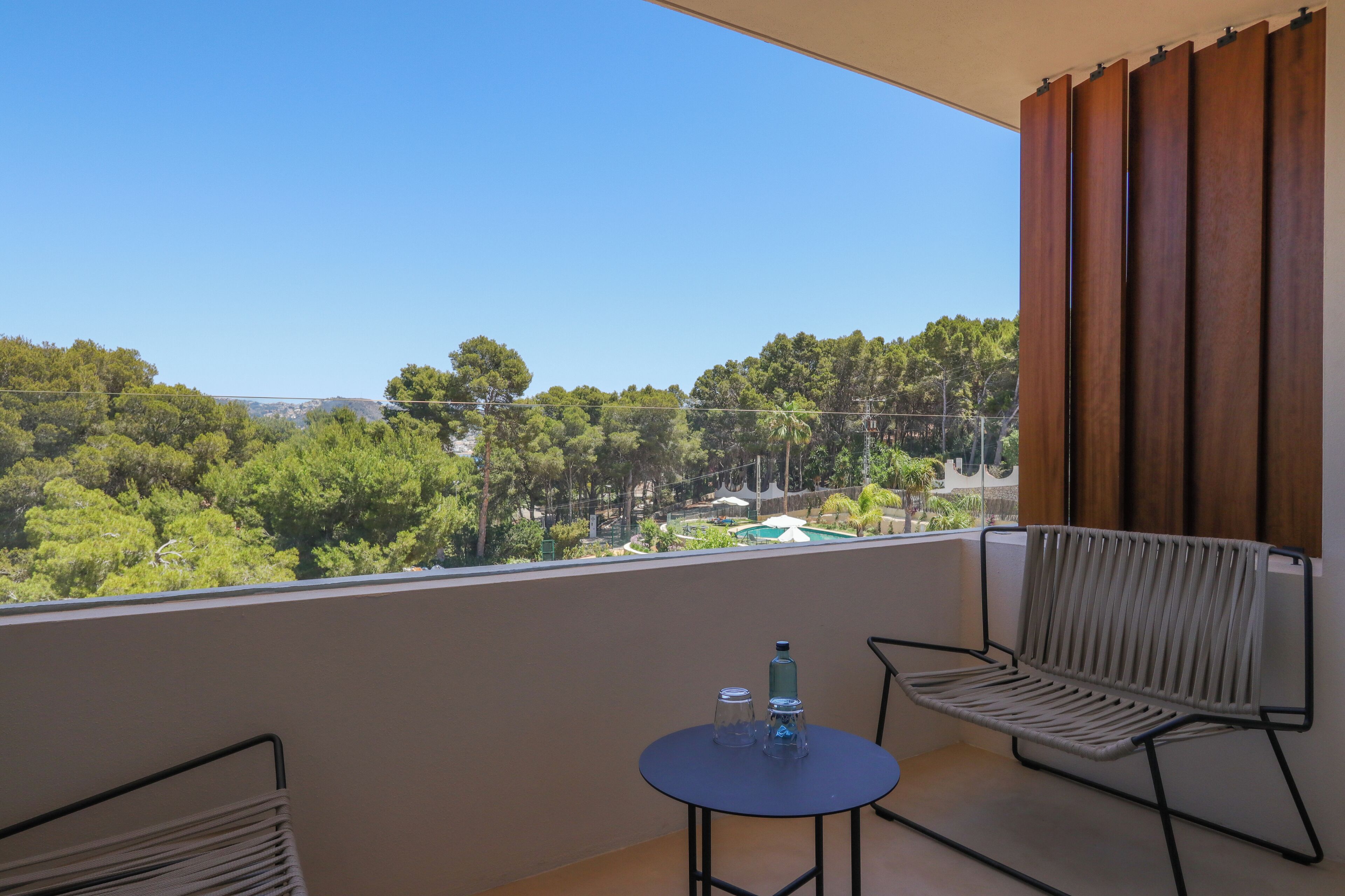 Een balkon met uitzicht op een bos en gebouwen in de verte, een tafel met twee glazen en een fles, en twee stoelen.