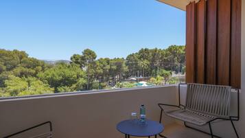 Een balkon met uitzicht op een bos en gebouwen in de verte, een tafel met twee glazen en een fles, en twee stoelen.