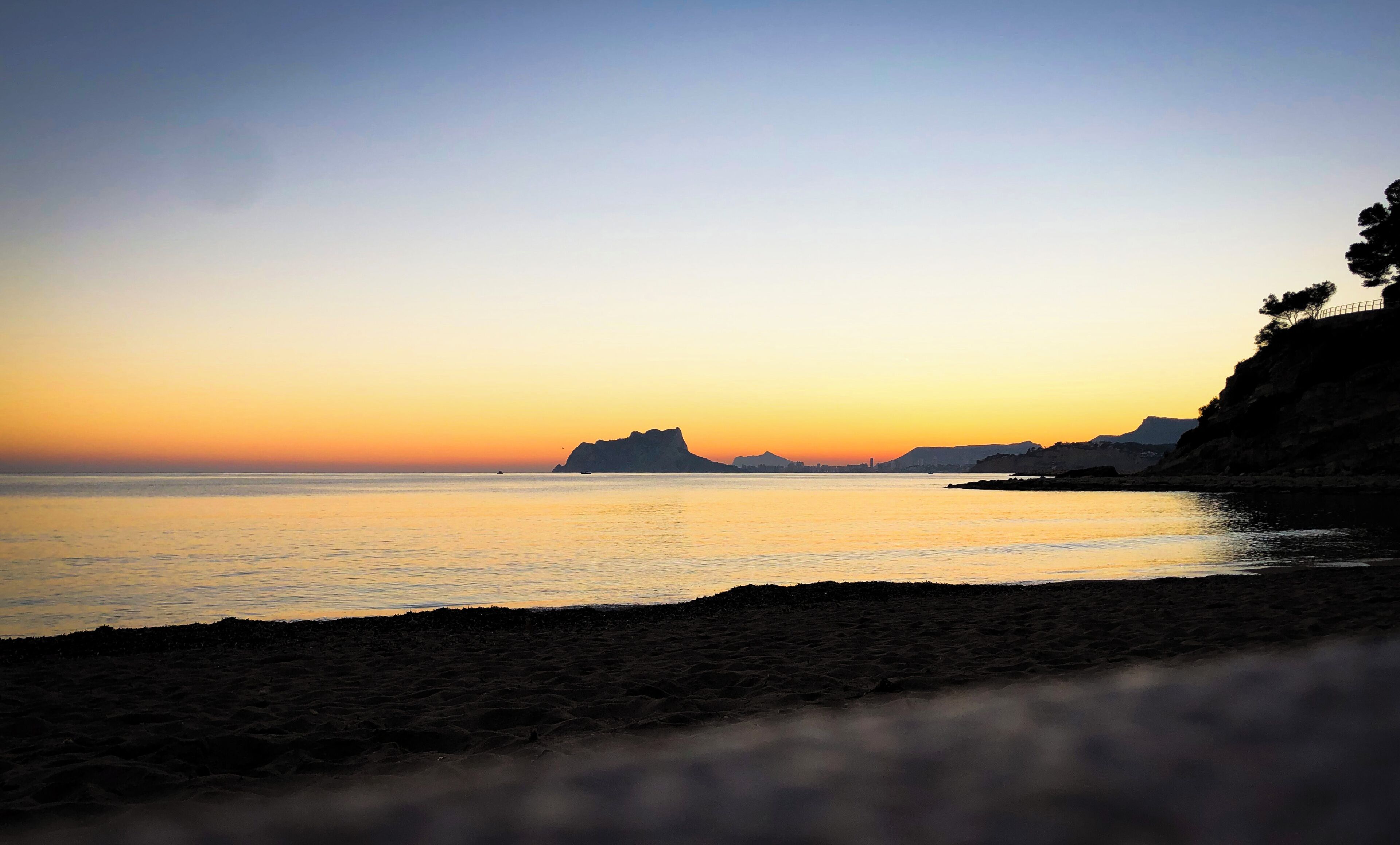 A beach at sunset with calm waters and distant landforms.
