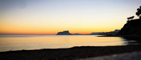 A beach at sunset with calm waters and distant landforms.