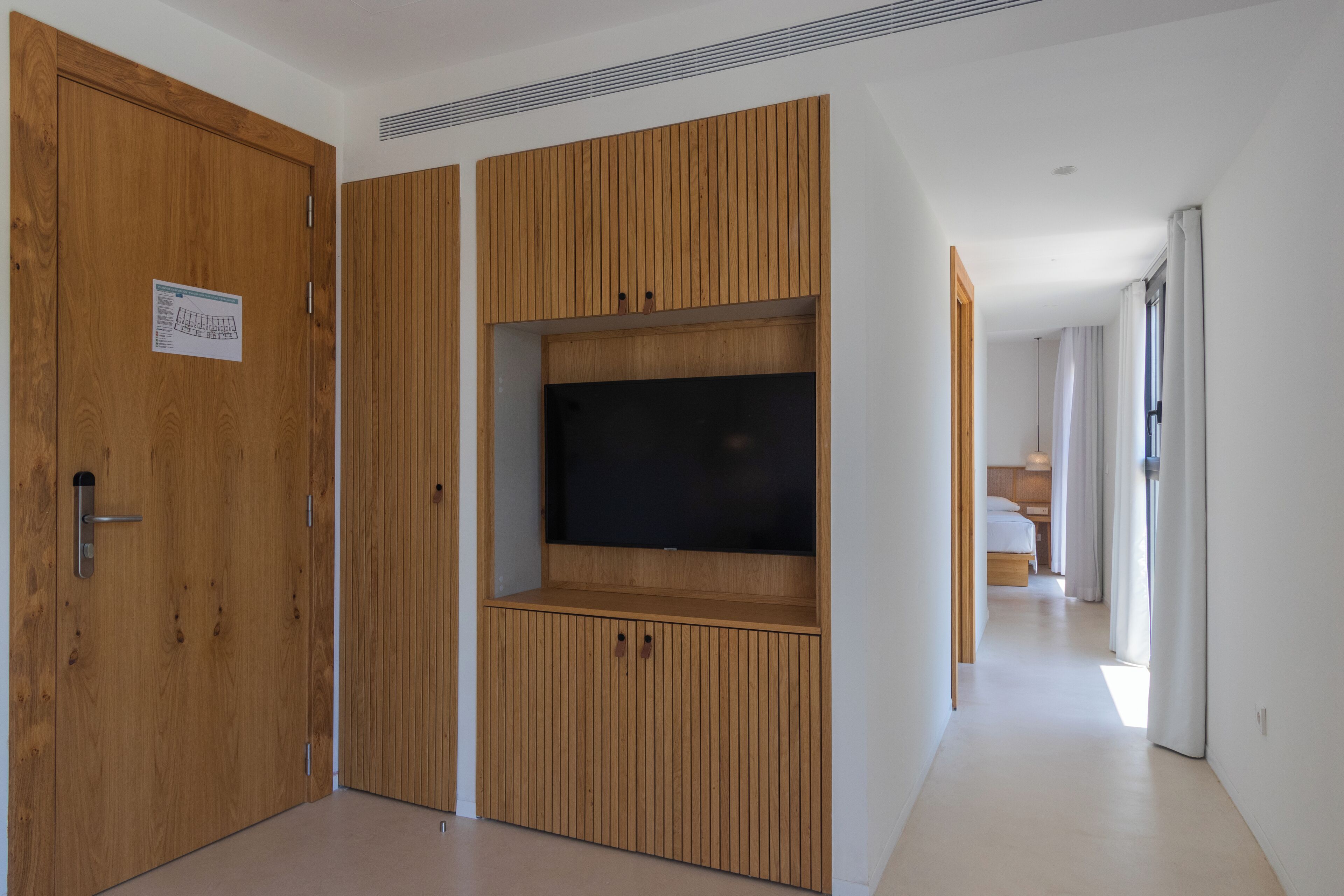 A hallway with a wooden door, a built-in TV, and a bed in the distance.