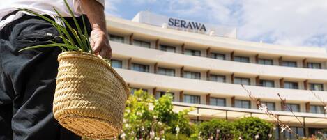 Una persona sostiene una cesta tejida con follaje, de pie frente a un edificio identificado como "SERAWA", con un jardín al fondo.