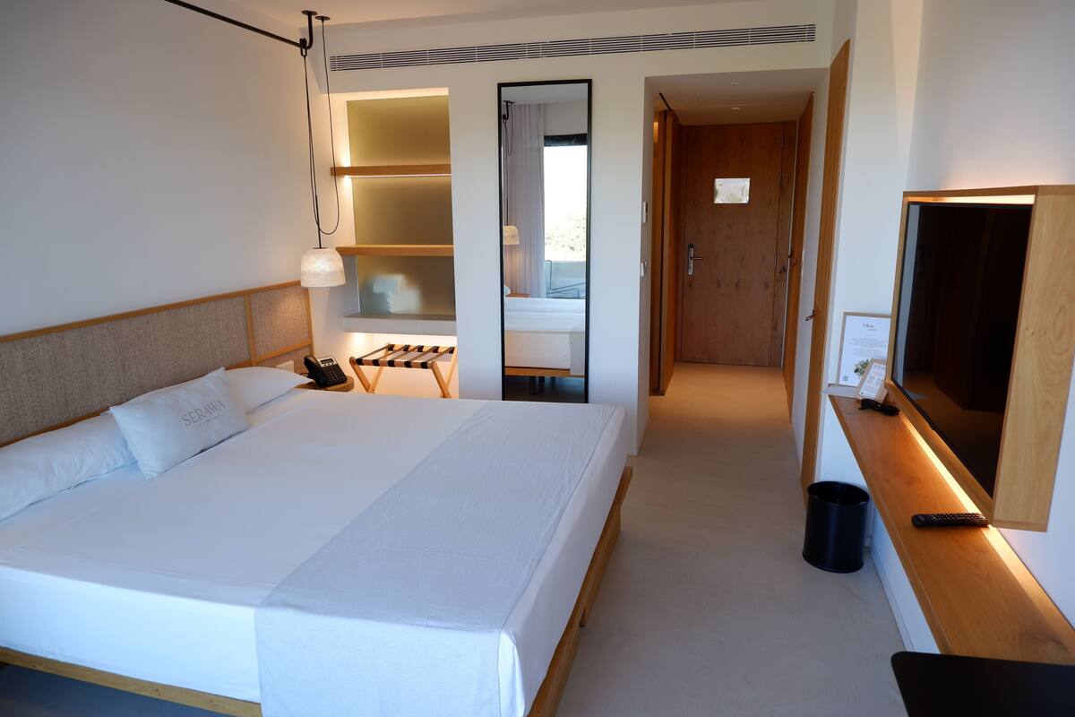Habitación de hotel con una cama grande, mesita de noche, lámpara y espejo. Se observa una puerta de madera y un pequeño cesto de basura.