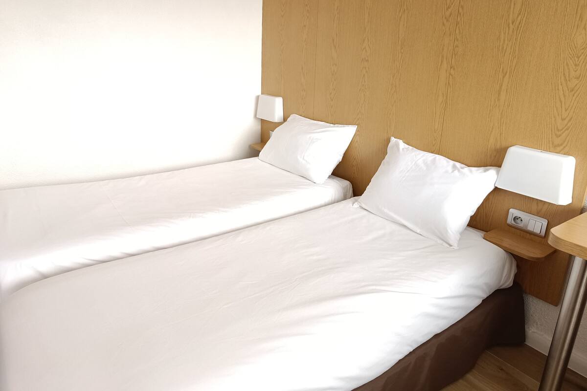 A hotel room with two single beds, wooden headboards, white bedding, and bedside tables with lamps.