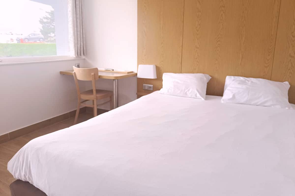A hotel room with a large bed, a wooden headboard, a desk with a chair, and a window with curtains.