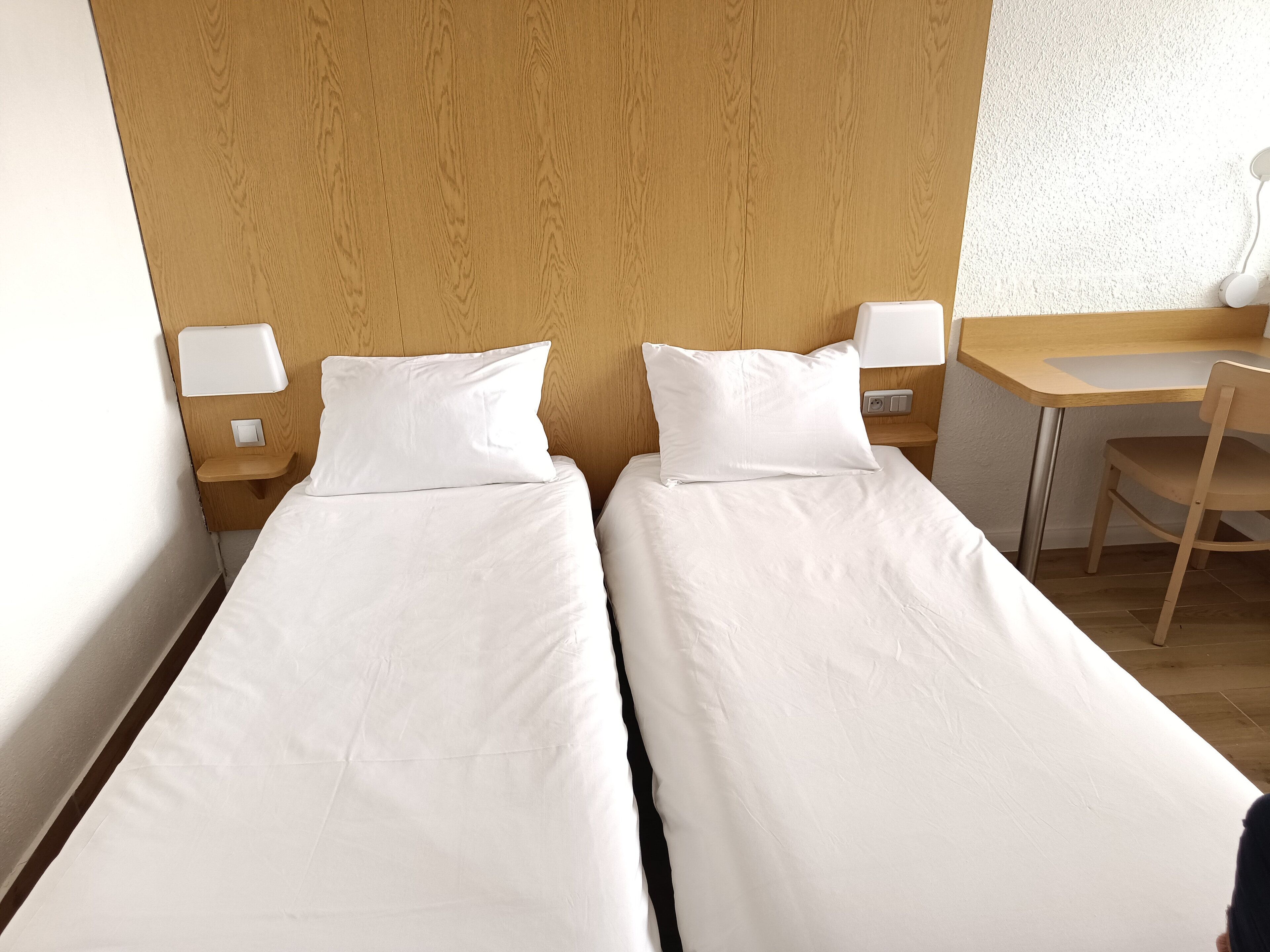 Habitación doble con dos camas individuales, cabecera de madera, escritorio, silla y mesita de noche con lámpara.